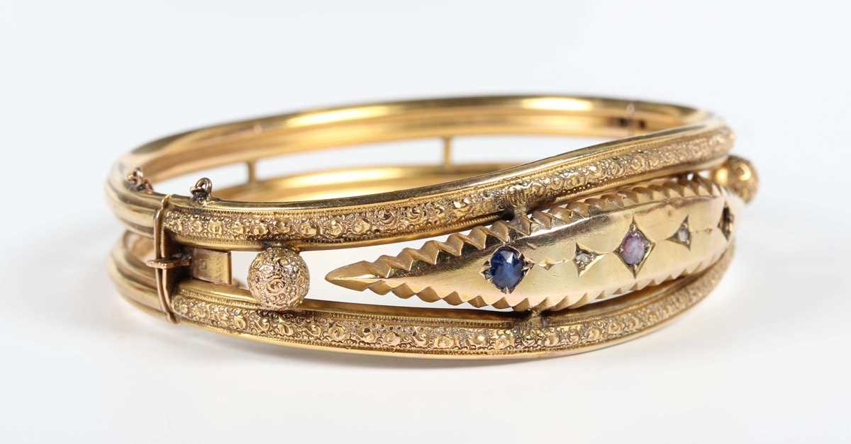 An Edwardian 9ct gold, diamond, blue gem and mauve paste set oval hinged bangle, decorated with