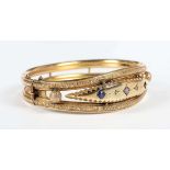 An Edwardian 9ct gold, diamond, blue gem and mauve paste set oval hinged bangle, decorated with