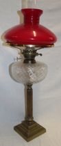 A brass oil lamp with fluted column and clear glass reservoir supporting a red opaque glass shade