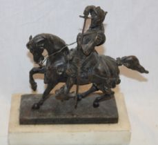 A bronze figure of a knight on horseback drawing a sword on a white marble base,