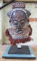 An African carved wood mask painted and decorated with shells, on stand, height 61cm.
