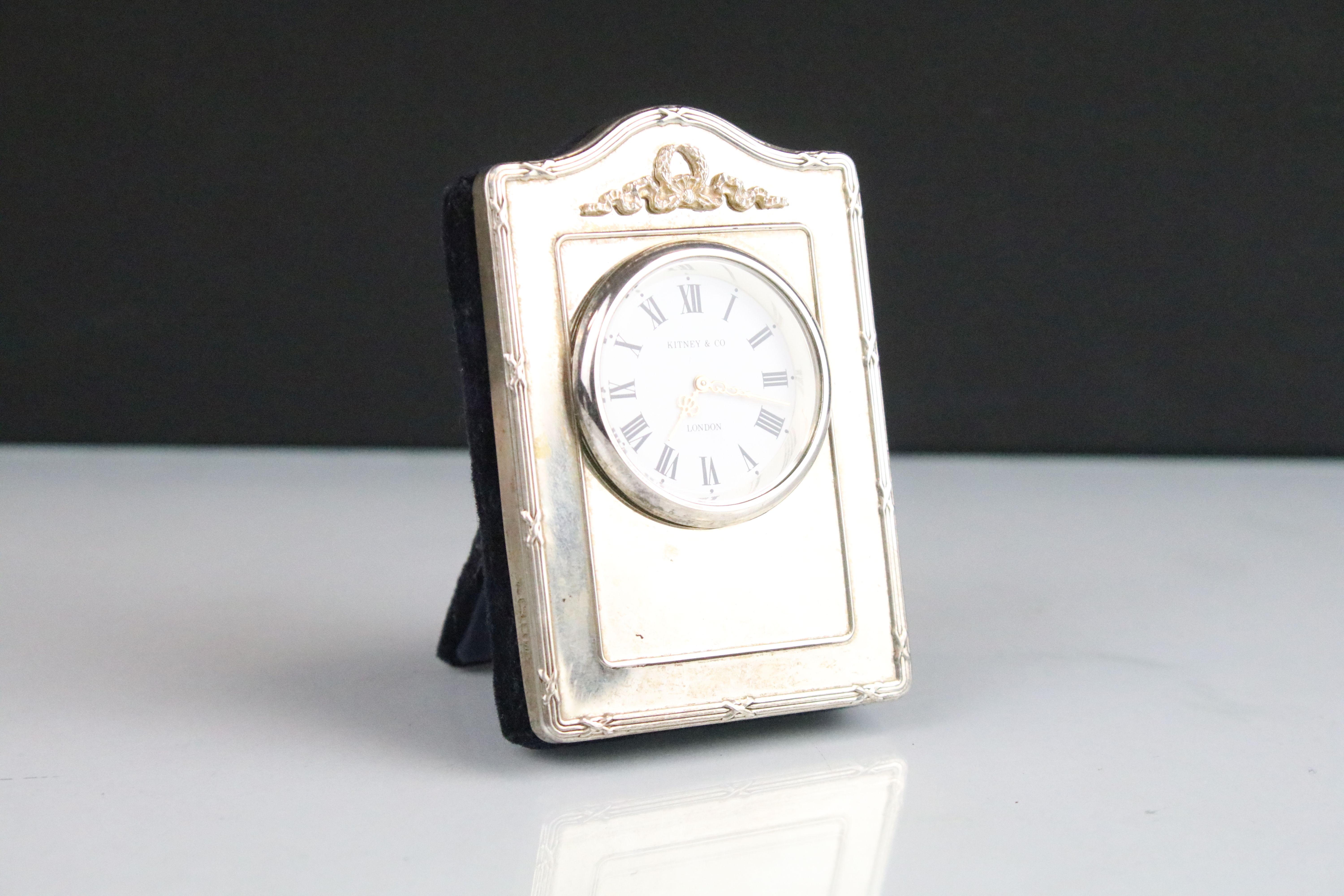 Kitney & Co silver fronted easel back clock timepiece having a round face with roman numerals to the