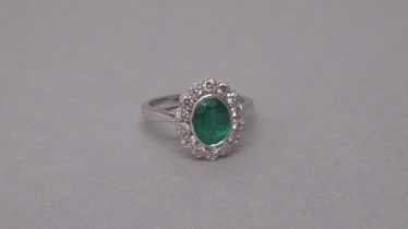 An 18ct white gold ring rubover-set with an oval emerald and a halo of round brilliant cut diamonds,
