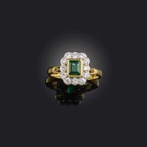 An emerald and diamond ring, of cluster design, set with a step-cut emerald within a border of