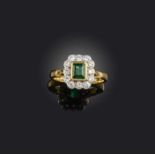 An emerald and diamond ring, of cluster design, set with a step-cut emerald within a border of