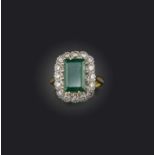 An emerald and diamond ring, of cluster design, claw-set with a step-cut emerald weighing