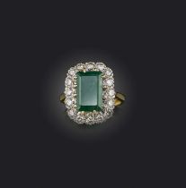 An emerald and diamond ring, of cluster design, claw-set with a step-cut emerald weighing