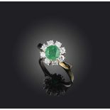 An emerald and diamond ring, of cluster design, set with a circular-cut emerald within a border of