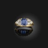 A sapphire and diamond ring, set with an oval sapphire, between shoulders set with circular-cut