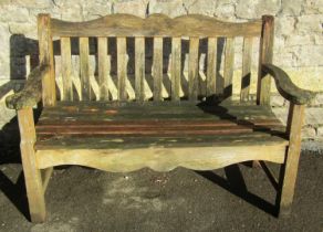 A weathered teak two seat garden bench with slatted seat and back beneath a shaped rail with