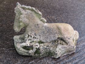 A weathered cast composition stone garden ornament in the form of a recumbent horse set on a