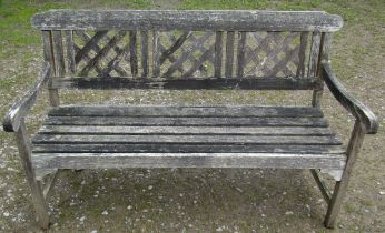 A weathered three seat garden bench with slatted seat and lattice panelled back beneath a shaped