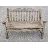 A Bridgman two seat weathered teak garden bench with slatted seat and back beneath a shaped rail
