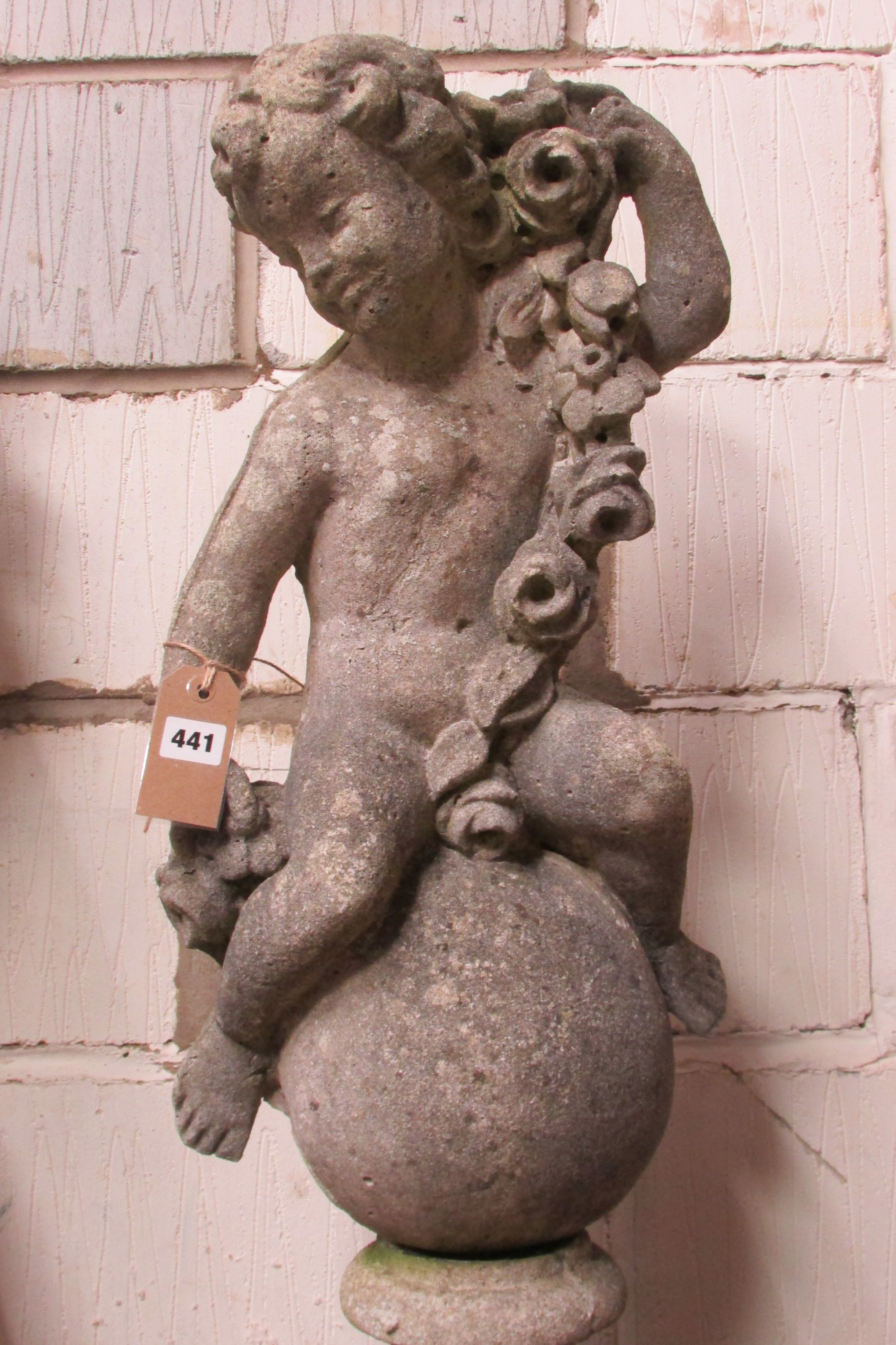 A weathered cast composition stone cherub astride a sphere clutching a floral garland raised on a - Image 2 of 3