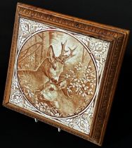 A decorative ceramic tile mounted in a wooden frame as a trivet, depicting a pair of roe deer in