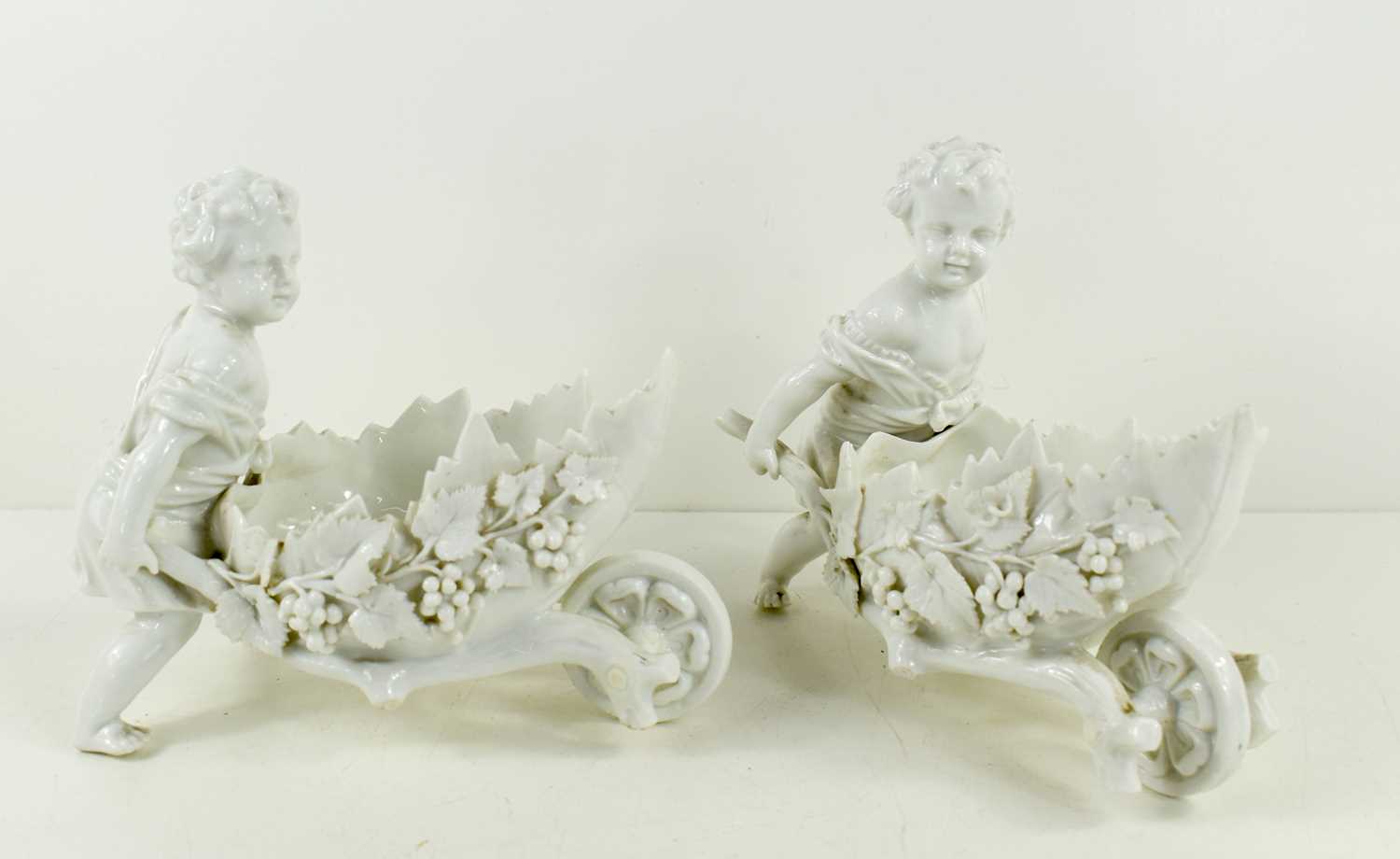 A pair of blanc de chine putti figurines, pushing leaf form wheel barrows, likely Triebner Ens &