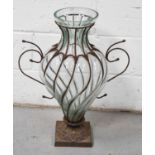A large glass and iron pedestal vase, of wrythen form, the hand blown glass with intricate iron