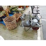 Metal wheel barrow containing galvanised watering cans and two wicker baskets