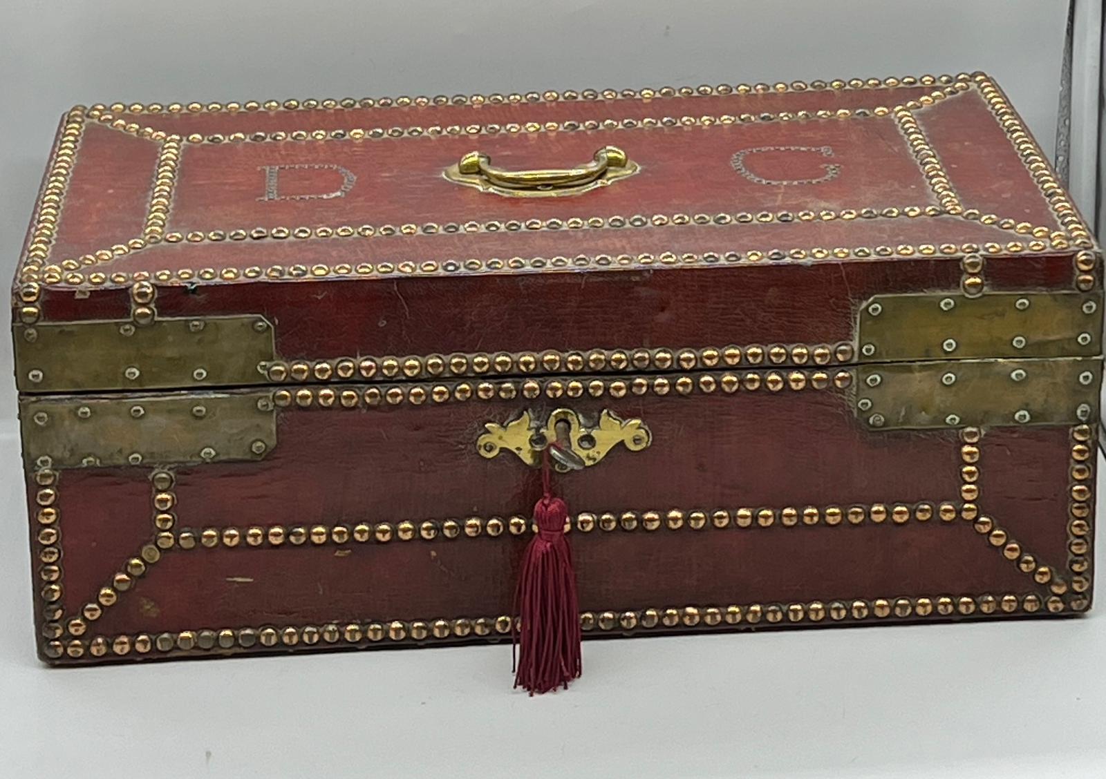 A red leather deed box decorated with studs brass plate corners. The initials DG are embedded with
