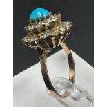 A turquoise central stone ring surrounded by clear stones on gold metal setting.