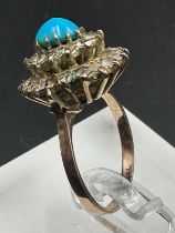 A turquoise central stone ring surrounded by clear stones on gold metal setting.