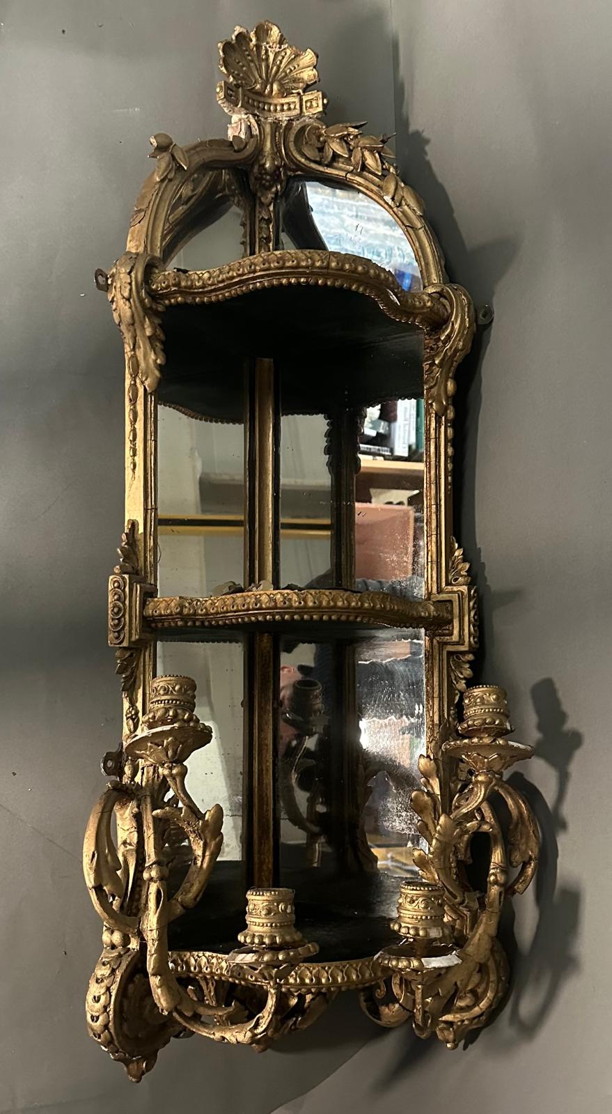 A gilt framed shelved corner mirror or girondele with two arm candle holders, two shelves and ornate
