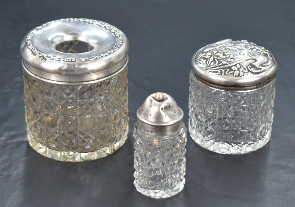A late Edwardian silver capped cut glass cosmetic container, having an embossed floral cap, along