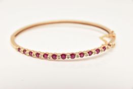 A YELLOW METAL GEM SET HINGED BANGLE, alternating circular cut rubies interspaced with collet set