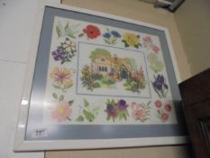 A framed and glazed tapestry/sampler of a cottage surrounded by flowers.
