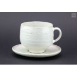 † LUCIE RIE (1902-1995); a stoneware cup and saucer covered in mottled and pitted white glaze,